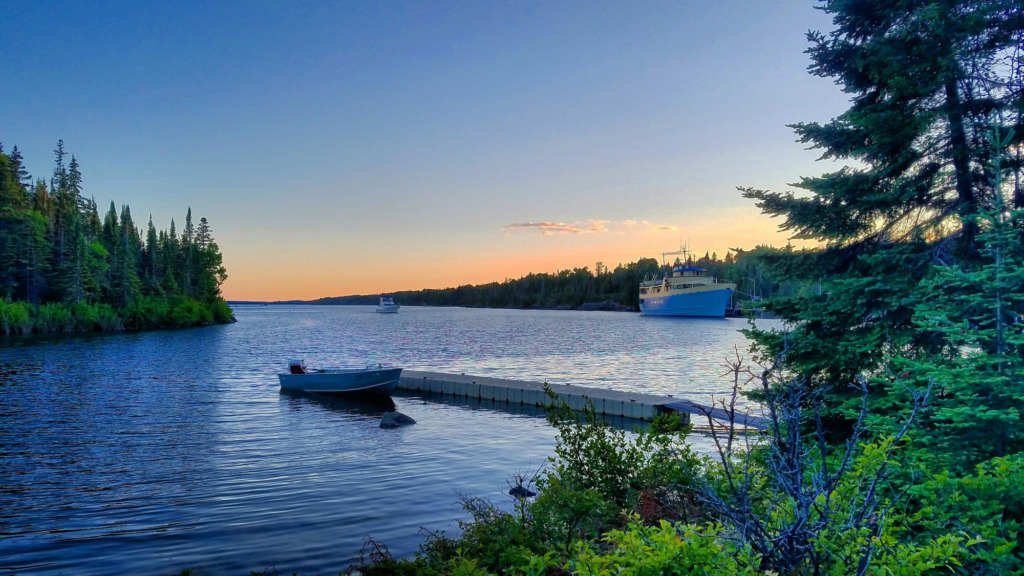 A large blue boat is in the far right of the frame with a simple pinkish sunset behind it. In the foreground is a dock with a small boat with trees and shrubs on both sides of the frame.